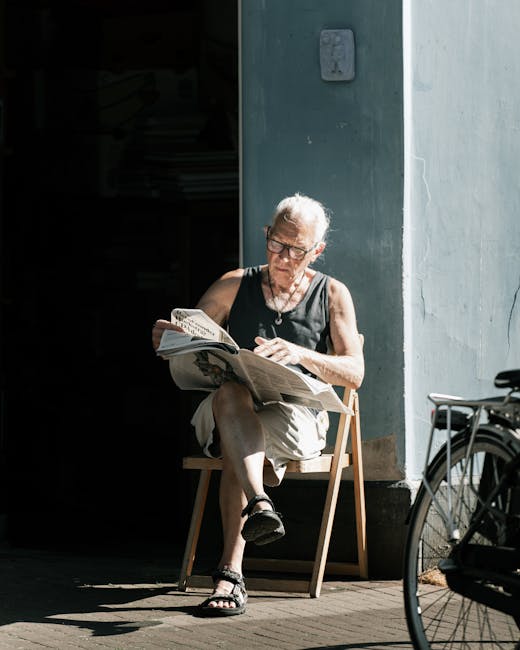 A senior woman with short grey hair, wearing glasses, a sleeveless black top, beige shorts, and black sandals, is seated on a wooden chair outside a building near a doorway. She is reading a newspaper