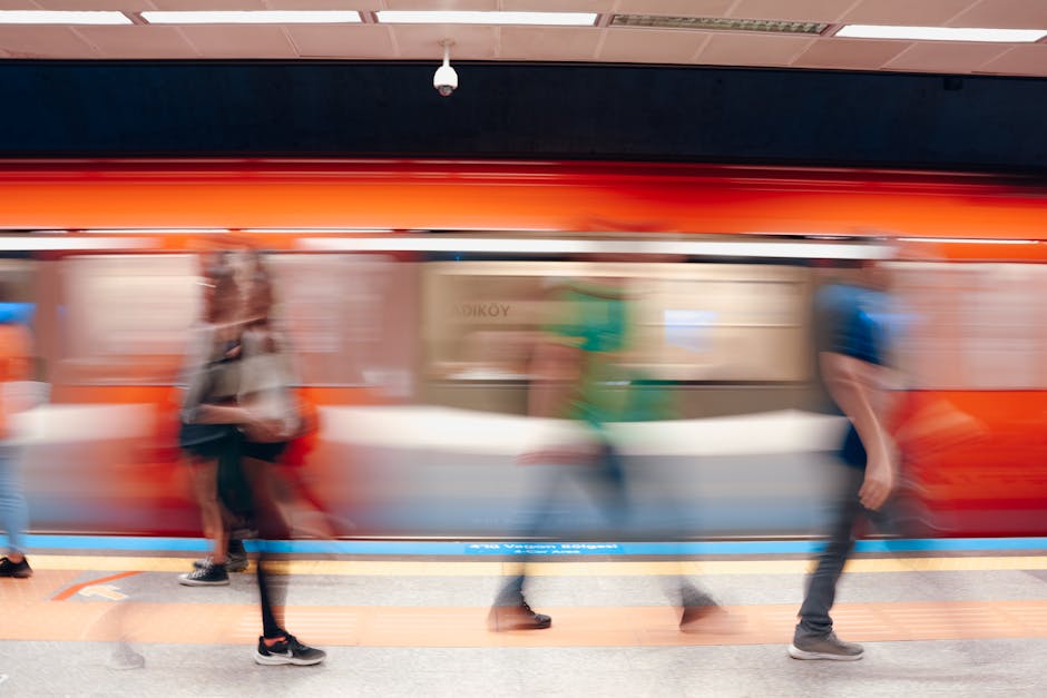 Inside a London underground station, three individuals are waiting on the platform for a train, which is in motion and slightly blurred as it departs. One man with a short haircut, wearing a white pol